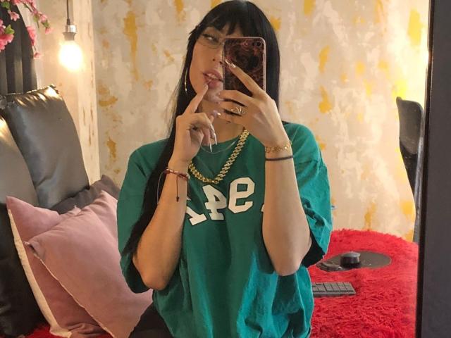 Violeta Parr, con cabello negro largo, camisa verde, collar dorado, dedo en la boca, sosteniendo un teléfono, alfombra roja, almohadas rosas, sofá de cuero negro, alfombra roja y pulsera roja..