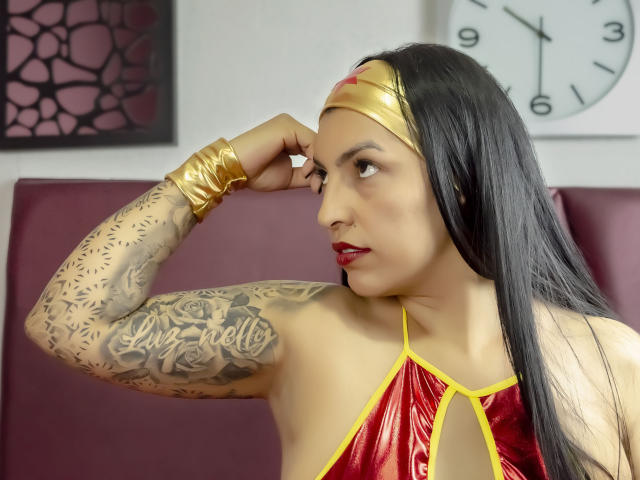 JanetteRider, avec ses longs cheveux noirs, son haut rouge brillant, son bandeau doré et ses tatouages sur les bras, lève son bras, exhibant sa poitrine et son bracelet en or. Elle porte du rouge à lèvres rouge et un anneau au nez..