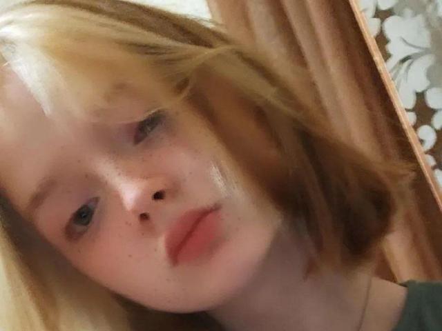 Das Gesicht von LaPochka ist im Fokus, mit blonden Haaren, Sommersprossen und leicht geöffneten, rosafarbenen Lippen. Sie hat einen weichen, leicht geröteten Teint mit blauen Augen und einem neutralen Ausdruck..