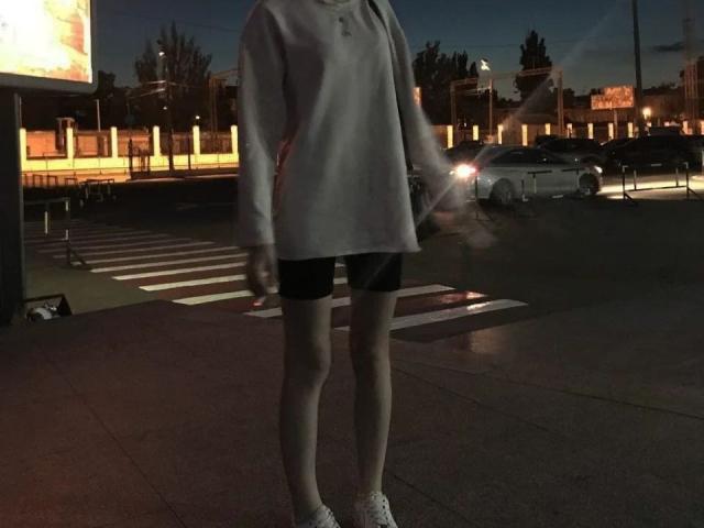 LaPochka with pale skin, wearing a loose gray sweater, black shorts, and white sneakers, standing in a dimly lit urban night scene, head out of frame.