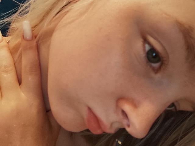 LaPochka's face, with blonde hair, slight freckles, and a pouty expression, has a hand gently touching her neck. Her green eyes are slightly upturned. Her nipples are not visible.