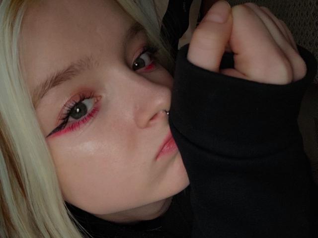 LaPochka, com pele clara, cabelo loiro, olhos verdes, delineador rosa, piercing no nariz, braço tatuado, segurando o braço tatuado, lábios levemente franzidos, close-up..