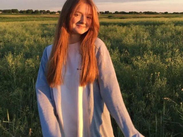 LaPochka, com longos cabelos ruivos, sorrindo, vestindo uma camisa azul clara por cima de uma camiseta branca, em pé em um campo gramado ensolarado, com as mãos relaxadas ao lado do corpo..