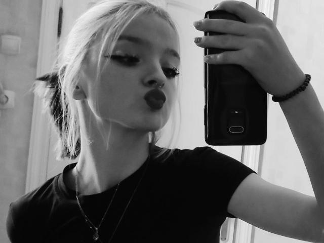 Selfie en blanco y negro de LaPochka con cabello rubio, piercing en la nariz y lápiz labial oscuro, haciendo pucheros, vistiendo una camisa negra, sosteniendo un teléfono y una pulsera, en un espejo..