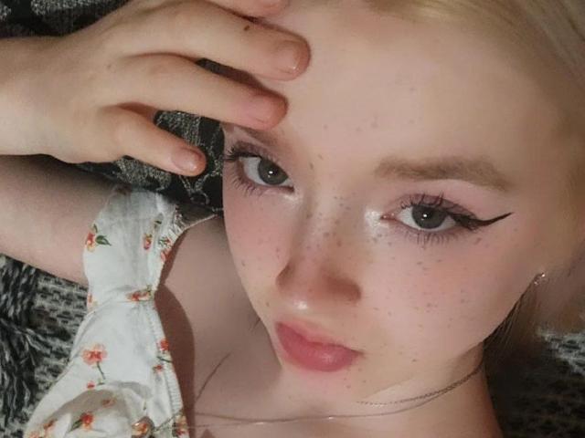 Le visage de LaPochka, avec ses cheveux blonds, son eye-liner rose, ses taches de rousseur et une main sur son front. Elle porte une robe à fleurs, regarde vers le haut avec les lèvres légèrement entrouvertes et d'une couleur rose..