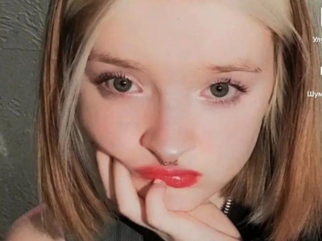 O rosto de LaPochka, com cabelo loiro, olhos verdes, lábios vermelhos, piercing no nariz e a mão perto da boca, exibindo lábios brilhantes e uma expressão fofa e emburrada..