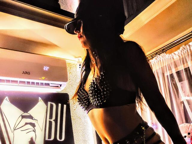 CarinFrancy, con lunghi capelli scuri, indossa un reggiseno nero con borchie, un berretto nero e occhiali da sole, ed è in piedi in una stanza poco illuminata, illuminata da una luce calda..