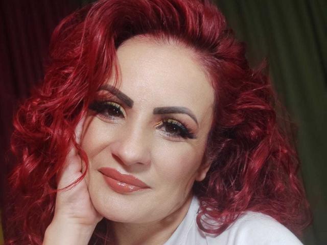 Karina Molina mit roten, lockigen Haaren, grünem und gelbem Lidschatten, dicken Augenbrauen und pinkem Lippenstift, die ihr Kinn in die Hand legt und mit einem leichten Lächeln in die Kamera schaut..