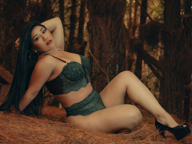 KhloeLawson with long black hair, wearing green lace bra and panties, black heels, and hoop earrings, sitting on pine needles, her hand behind her head, her ass prominent.