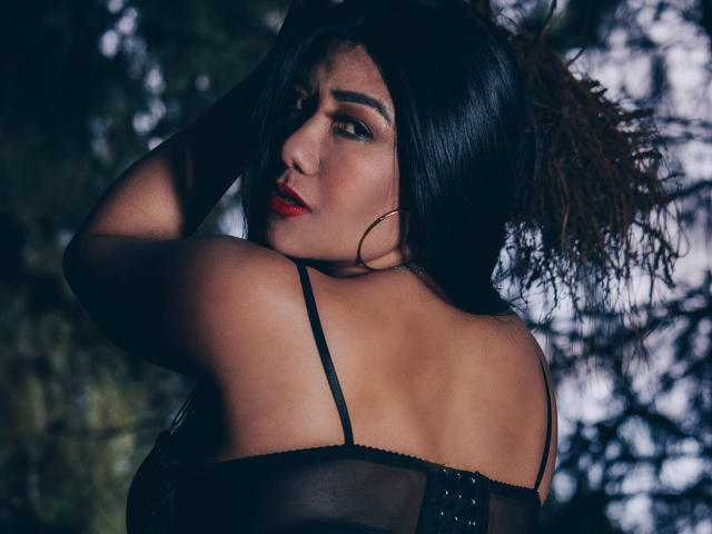 KhloeLawson with dark hair, red lipstick, black sheer top, looking back, hand in hair, showing her bare shoulders and upper back, looking sexy and confident.