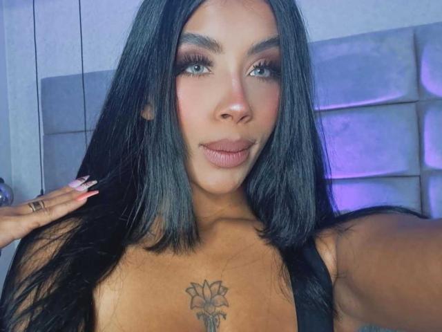 FiorellaBossh, com cabelos pretos longos, sobrancelhas grossas e uma tatuagem de flor no peito, vestindo uma blusa preta, olhando para a câmera com um leve sorriso..