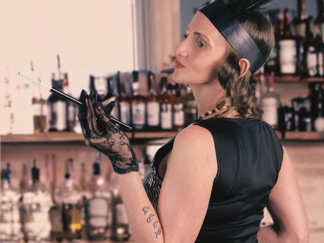 RubiAnderson with black lace gloves, holding a cigarette, wearing a black dress and metallic headband, showing tattoos on her right arm, looking over her shoulder.