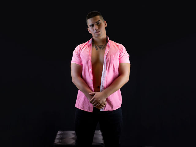 KingaNDher, um homem musculoso com cabelo preto curto, vestindo uma camisa rosa desabotoada que revela uma tatuagem no peito, calças pretas, com as mãos unidas na frente, em pé contra um fundo escuro..