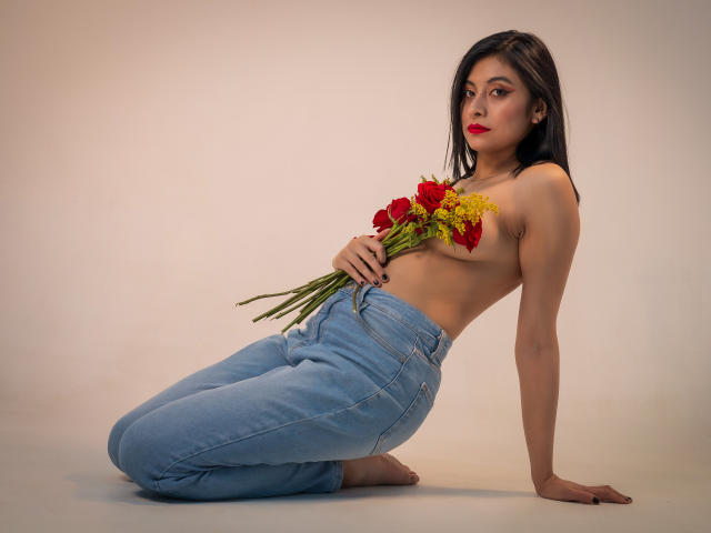 Samanthaa Smith está desnuda de arriba, arrodillada, con jeans azules, sosteniendo rosas rojas y flores amarillas, cubriendo sus pechos con el ramo, tiene lápiz labial rojo y una expresión neutra..