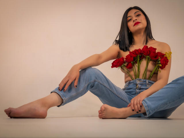 Samantha Smith, sin sostén, sentada con las piernas separadas, sosteniendo rosas rojas sobre sus pechos, vestida con jeans azules, descalza, con esmalte de uñas negro, lápiz labial rojo, cabello negro, segura de sí misma, sexy..