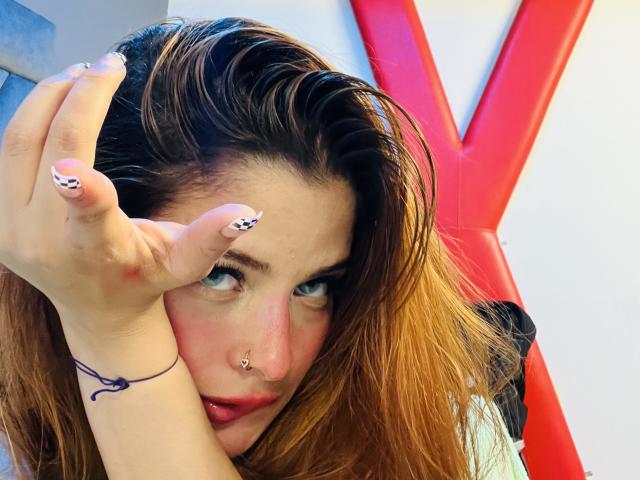 ShantalSummer tiene el cabello castaño despeinado, ojos azules y un piercing en la nariz, y hace un gesto con la mano formando una "V", mostrando uñas manicuradas en blanco y negro y una pulsera morada..