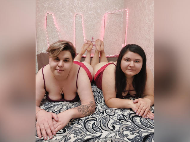 BeckyAndEllen, entrambe in lingerie rossa, sono sdraiate su un letto con lenzuola a motivi bianchi e neri, mostrando i loro seni e glutei, con i piedi sollevati, e delle lettere al neon sullo sfondo..