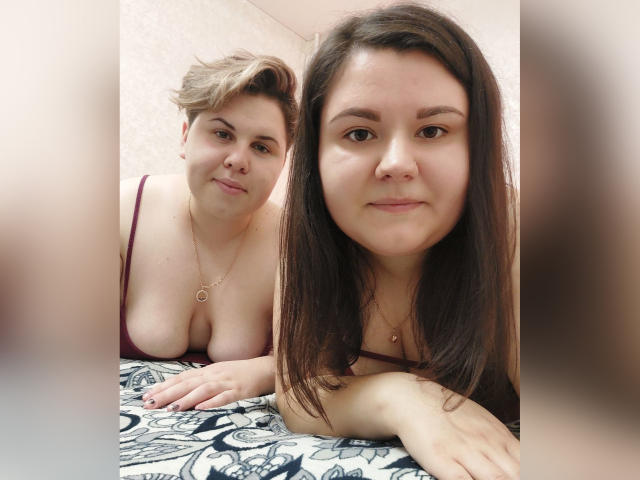 BeckyAndEllen, due donne, una con i capelli biondi corti, un seno abbondante e un reggiseno rosso, e l'altra con i capelli castani lunghi, sdraiate su un letto a motivi, entrambe sorridenti..