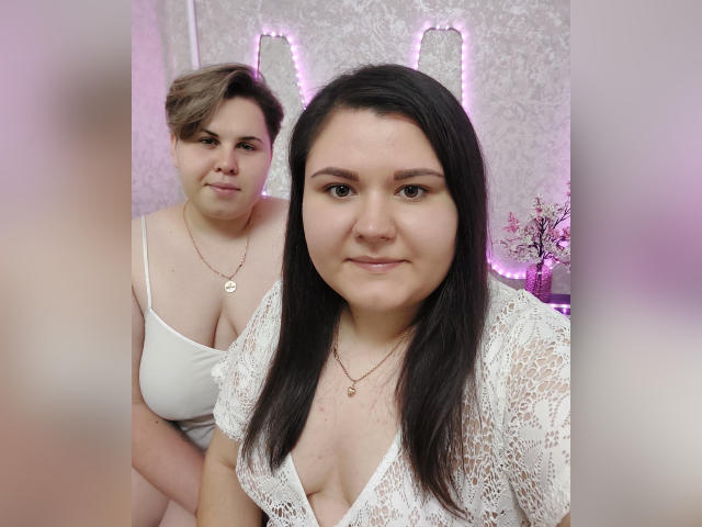 BeckyAndEllen with short brown hair and small tits in a white top, standing behind BeckyAndEllen with long black hair and large tits in a lace top.