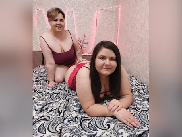 BeckyAndEllen with tits in a red bra, kneeling, smiling, tattoo on arm, behind Ellen, who has black hair, red panties, lying on a patterned bed, smiling.