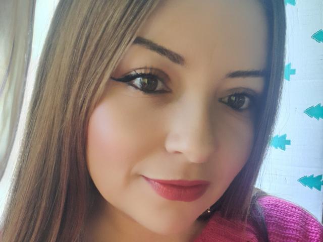 NatalyHami's face with long brown hair, green eyes, black eyeliner, and pink lipstick, smiling softly. She's wearing a pink sweater. Her ass is not visible.