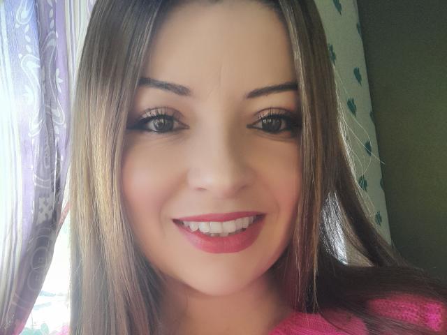 NatalyHami ha lunghi capelli castani, sopracciglia scure, occhi verdi e rossetto rosso, e sorride mostrando denti bianchi. Indossa una maglietta rosa, e la foto è un selfie..