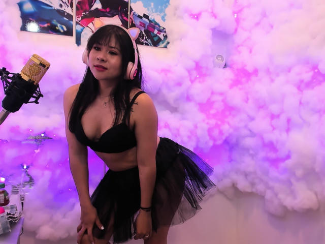 LourrieAnn, usando fones de ouvido rosa, um sutiã preto e uma saia preta transparente, inclinada para frente com seios pequenos, cabelo preto e um microfone em um quarto rosa cheio de fumaça..