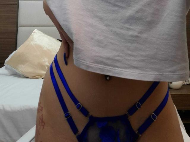 A parte inferior da barriga de ChanelDeRosee, vestindo lingerie azul com alças finas, com um piercing visível no umbigo, uma tatuagem na parte direita da virilha e uma camisa branca que cobre parcialmente o corpo..