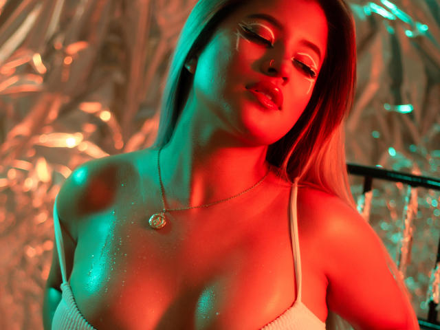 MegannSweettx mit leuchtendem roten und grünem Licht, glänzender Haut, weißem Top mit Spaghetti-Trägern, einer kleinen, runden Halskette, geschlossenen Augen, leicht geöffneten Lippen, sichtbarem Ausschnitt, langen blonden Haaren, entspanntem Gesichtsausdruck..