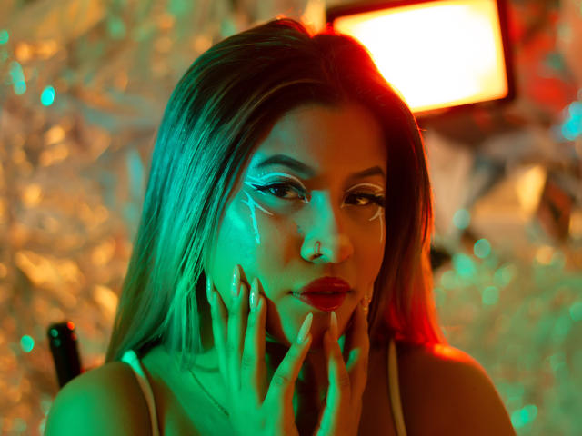 MegannSweettx avec un éclairage vert et rouge, les mains sur le visage, de longs cheveux blonds, un eye-liner dramatique, du rouge à lèvres rouge, regardant la caméra, un bokeh doux, une lumière douce en arrière-plan..