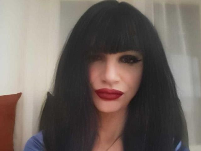 Juny, avec ses cheveux noirs, lisses et mi-longs, portant un rouge à lèvres rouge vif, a un maquillage des yeux foncé et saisissant, et regarde l'appareil photo avec un léger sourire confiant..