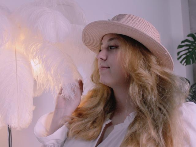 Jessika Daniels, con i capelli biondi mossi, indossa un cappello di paglia, una camicia bianca e guarda una lampada bianca e soffice. Il suo seno è parzialmente visibile. Luce naturale e delicata..