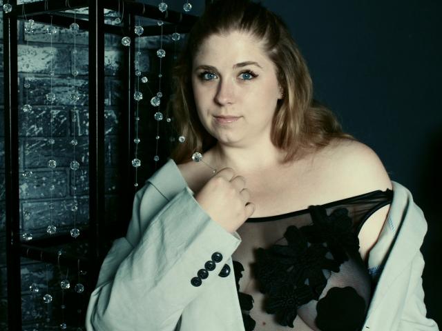 Jessika Daniels, aux yeux bleus, avec des cheveux bruns clairs, portant un haut noir transparent avec des motifs floraux, à épaules dénudées, une veste grise, tenant la veste, regardant la caméra, plan moyen, fond sombre..