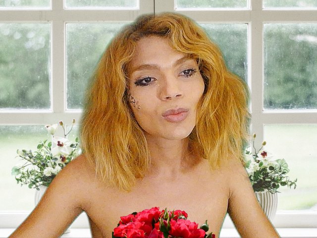 TatyLorenX con cabello amarillo-anaranjado, labios carnosos y hombros desnudos sosteniendo rosas rojas, de pie frente a una ventana con flores blancas a cada lado..