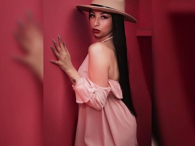 CamilleRios with long black hair, wearing a pink off-shoulder dress, beige hat, and red lipstick, touches a red wall with her right hand, showing a tattoo on her wrist.