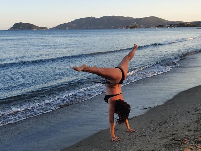 JolieXKitty is upside down on the beach, wearing a black bikini, with one leg raised high, her ass and tits visible, and her pussy slightly exposed.