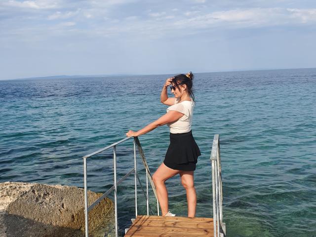 JolieXKitty está de pie en un muelle de madera, vestida con una blusa blanca y una falda negra, con la mano en su cabello castaño, mirando el océano azul. Se puede ver su trasero..