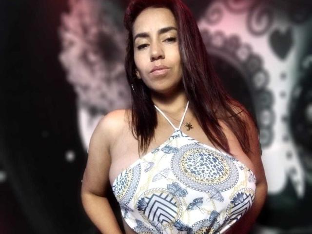 KatyKatt, avec ses cheveux brun foncé, portant un haut à encolure dégagée avec des motifs bleus et blancs, mettant en valeur ses seins volumineux et ronds, et affichant une expression confiante, légèrement boudeuse..