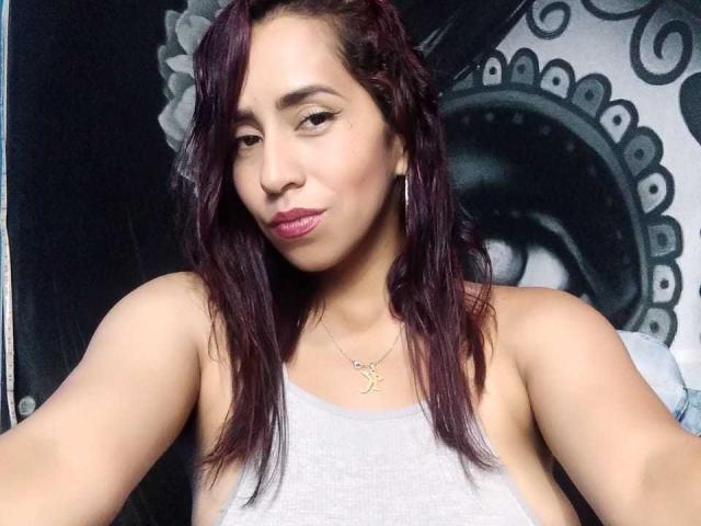 KatyKatt with dark purple hair, gray tank top, small gold necklace, pink lipstick, and slight smile, showing her bare shoulders and upper chest. Her tits are partially visible.