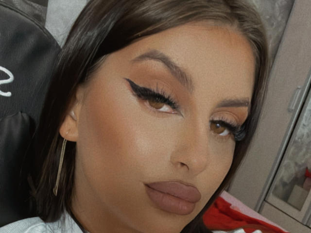 CassieSin has tan skin, brown eyes, and brown eyeliner with thick lashes. She has full brown lips and is wearing gold hoop earrings. Her brown hair is straight.