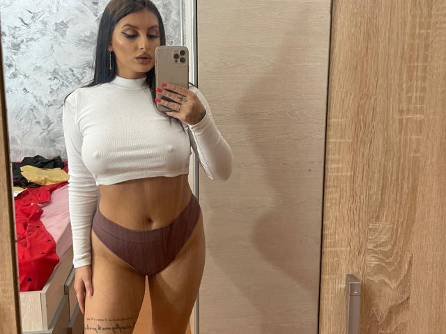 CassieSin, avec ses longs cheveux noirs, portant un crop top blanc et une culotte haute mauve, se tient devant un miroir, tenant un iPhone, et montrant ses seins et ses fesses..