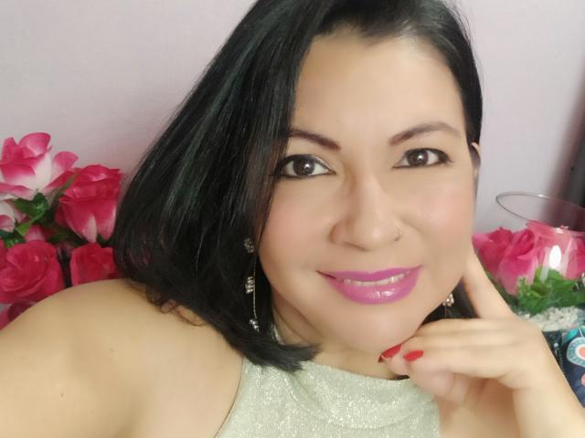 Amanda Kohler, com batom rosa brilhante, cabelo preto na altura dos ombros e unhas vermelhas, sorrindo, vestindo uma blusa brilhante, com um piercing no nariz e brincos, cercada por rosas cor-de-rosa..