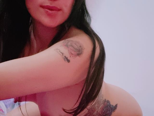 LindaSmith is nude, with long black hair, red lips, a tattoo on her upper arm, and another tattoo on her right side, showing her tits and ass.