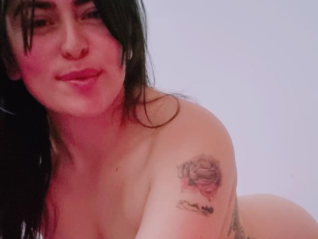 LindaSmith has dark hair, a nose ring, and a rose tattoo on her right shoulder. She's nude, with her tits and ass partially visible, smiling slightly.