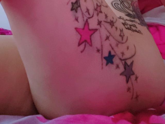 LindaSmith's butt tattoo with colorful stars and swirls, featuring pink, blue, and black stars, and a heart design, visible in a pink bedspread.