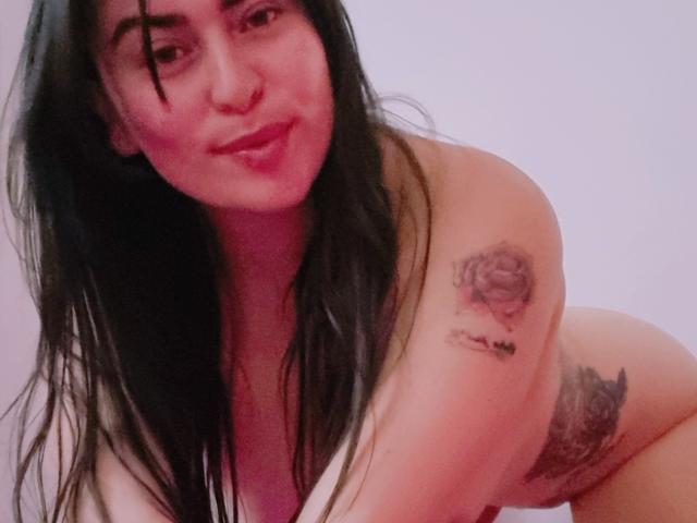 LindaSmith with long black hair, pouty lips, nose ring, and tattoos on her shoulder and side, leaning forward, topless, showing her tits. She's smiling at the camera.