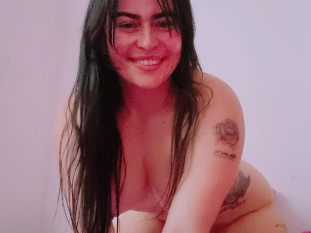 Nude LindaSmith with long black hair, smiling, leaning forward. She has tattoos on her right arm and left side, and she's showing her big tits and soft ass.