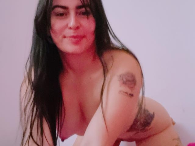 Nude LindaSmith with long black hair, medium tits, and tattoos on her right arm and left hip, leaning forward with a slight smile. Her skin is light with a pinkish hue.