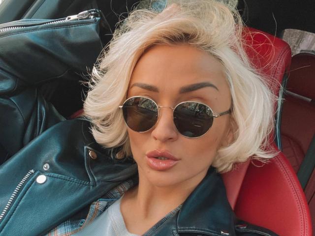 XhotLoli, avec ses cheveux blonds platine, ses lunettes de soleil rondes de type aviateur, ses lèvres roses brillantes et sa veste en cuir noir, assise sur un siège de voiture rouge, regardant directement l'appareil photo..