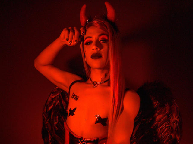 Alessandra Ferrer, con corna da diavolo rosse, rossetto nero e lunghi capelli biondi, indossa una collana di pelle e dei piccoli copri-capezzoli neri, ha delle ali e un tatuaggio sul petto. Luce rossa..